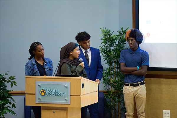 Image of four students standing on a stage while presenting their work.