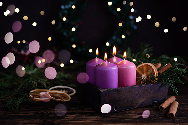 Four purple burning advent candles on a dark wooden background with festive lights.